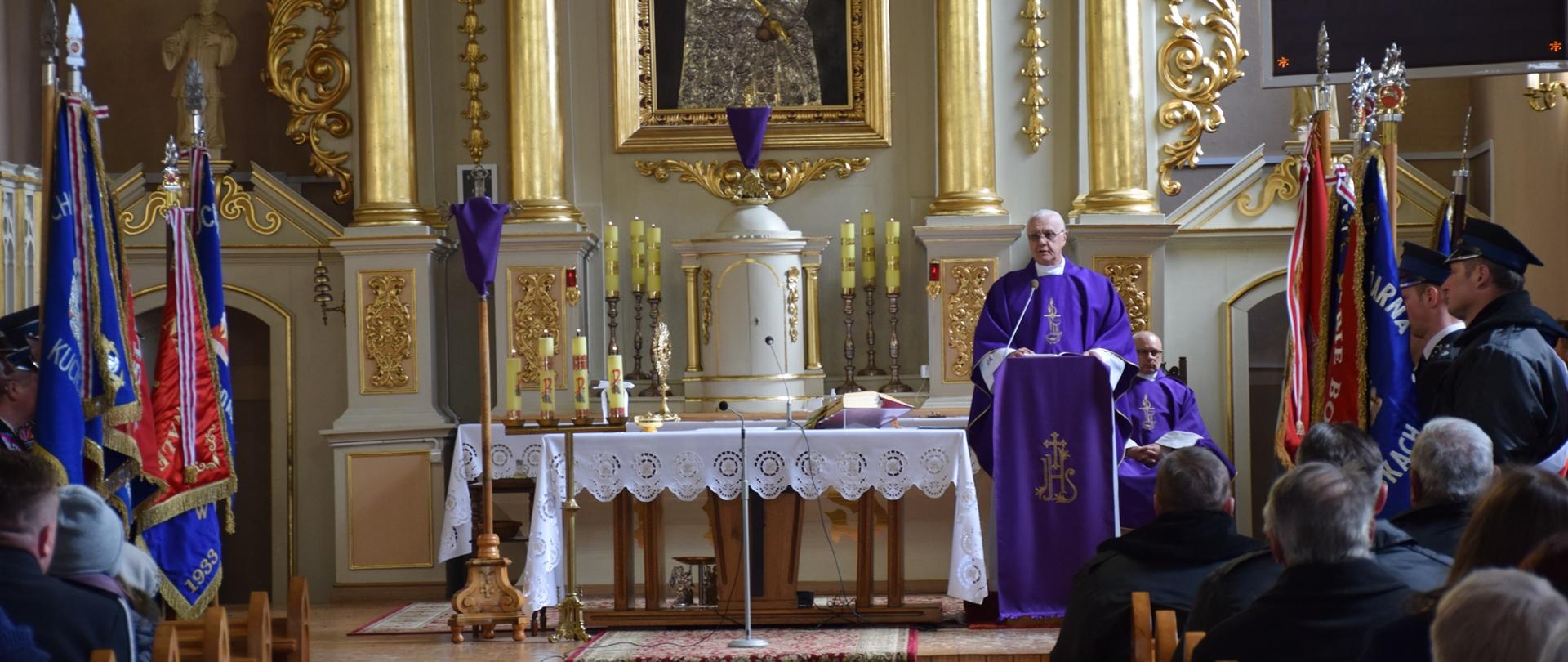 Ołtarz w Kościele, Na mównicy stoi ksiądz w szatach koloru fioletowego. Po bokach poczty sztandarowe.