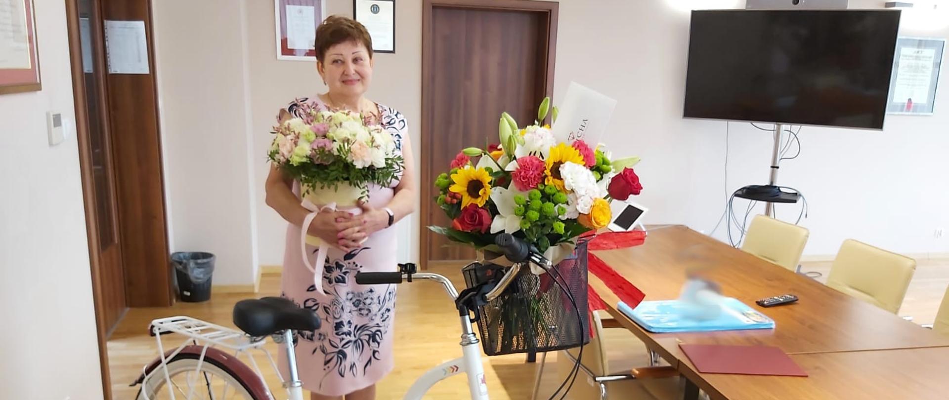 Osoba odchodząca na emeryturę stoi z kwiatami a przed nią stoi rower.