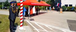 Fotografia przedstawia na pierwszym planie po lewej stronie poczet flagowy składający się z trzech strażaków. Na dalszym planie widać dwa namioty, pod którymi znajdują się zaproszeni goście. Po prawej stronie w oddali mównicę wraz z budynkiem Jednostki Ratowniczo Gaśniczej nr 2 Sosnowiec „Porąbka”.