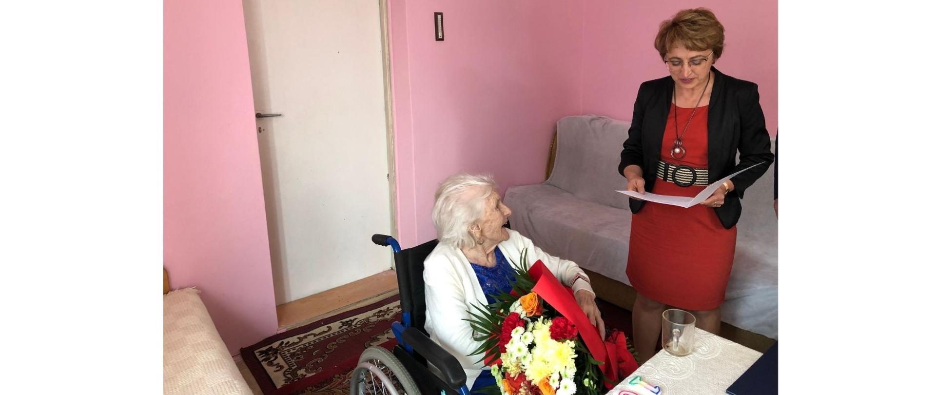 Na zdjęciu znajduje się starsza kobieta na wózku a po prawej stronie stoi kobieta w czerwonej sukience i odczytuje list