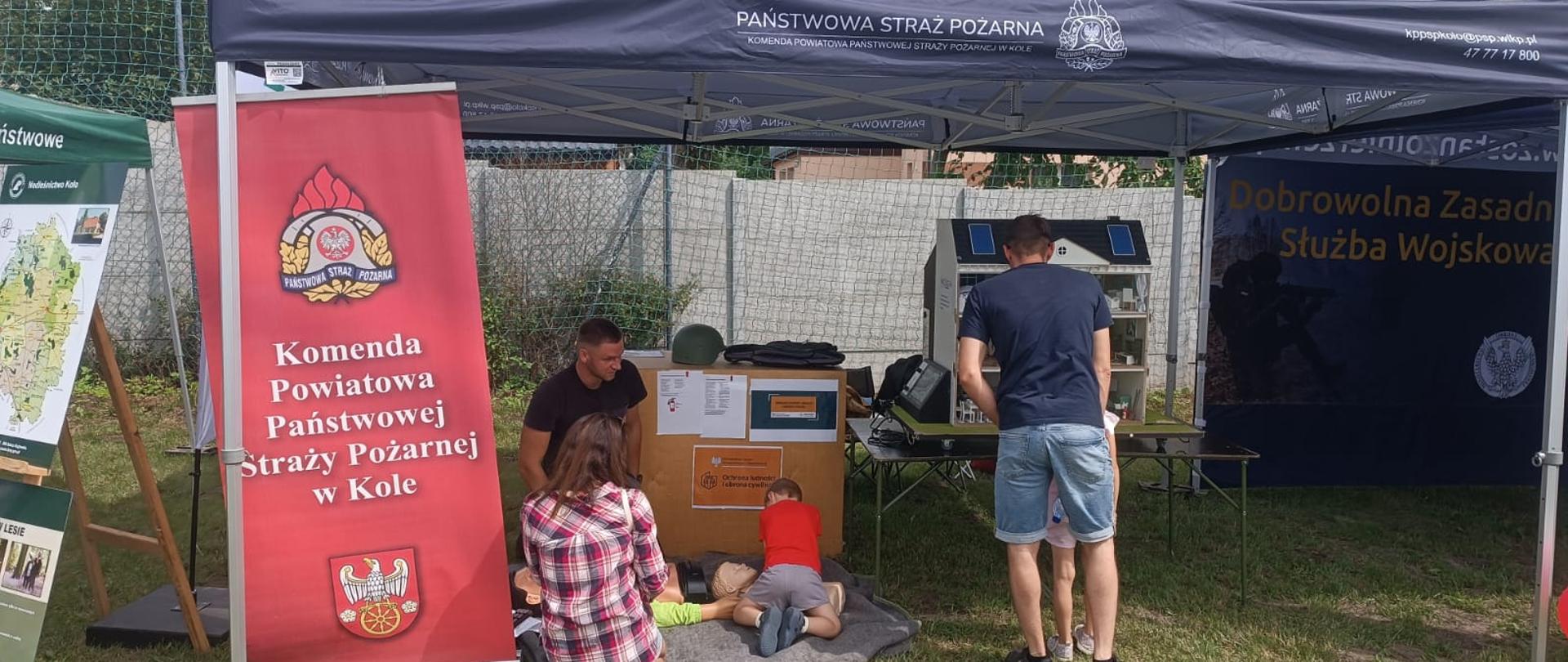 Stoisko Komendy Powiatowej Państwowej Straży Pożarnej w Kole podczas wydarzenia plenerowego, gdzie prowadzone są pokazy pierwszej pomocy dla dzieci.