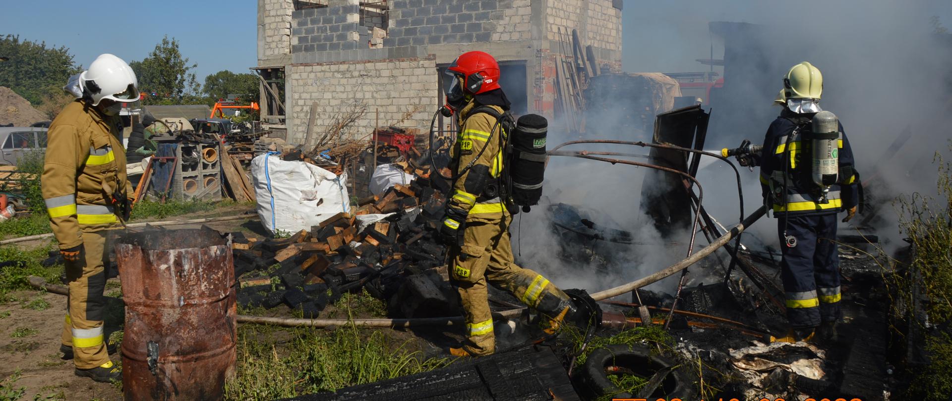 Pożar drewnianej szopy. Na pierwszym planie metalowa beczka, drewno w big-bagach oraz luzem i trzech strażaków prowadzących działania ratowniczo-gaśnicze. Z prawej strony widoczne jest niewielkie zadymienie. W tle budynek w surowym stanie. 