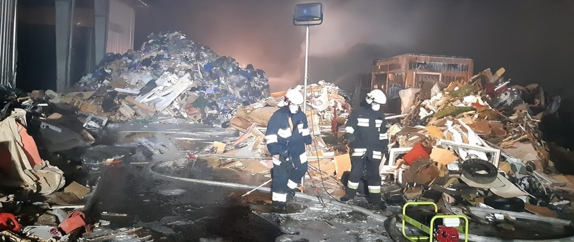 Strażacy OSP przeszukujący teren pożaru. Teren akcji oświetlony najaśnicą. Dookoła rozrzucone spalone rzeczy.