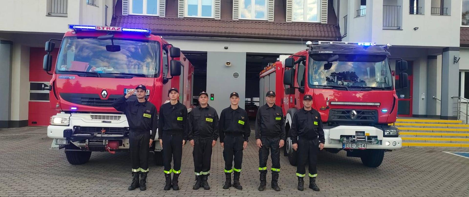 Na zdjęciu widać sześciu strażaków stojących w jednym szeregu oraz dwa samochody strażackie na placu przed budynkiem Komendy Powiatowej PSP w Łęczycy. Strażacy ubrani sa w czarne stroje z napisem straż oraz czapki z daszkiem. Jest widno.