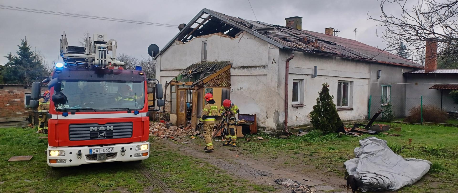 Wóz strażacki na prywatnej posesji po wybuchu gazu. po prawej stronnie zniszczony budynek z naruszoną konstrukcją dachu i wejściem do budynku