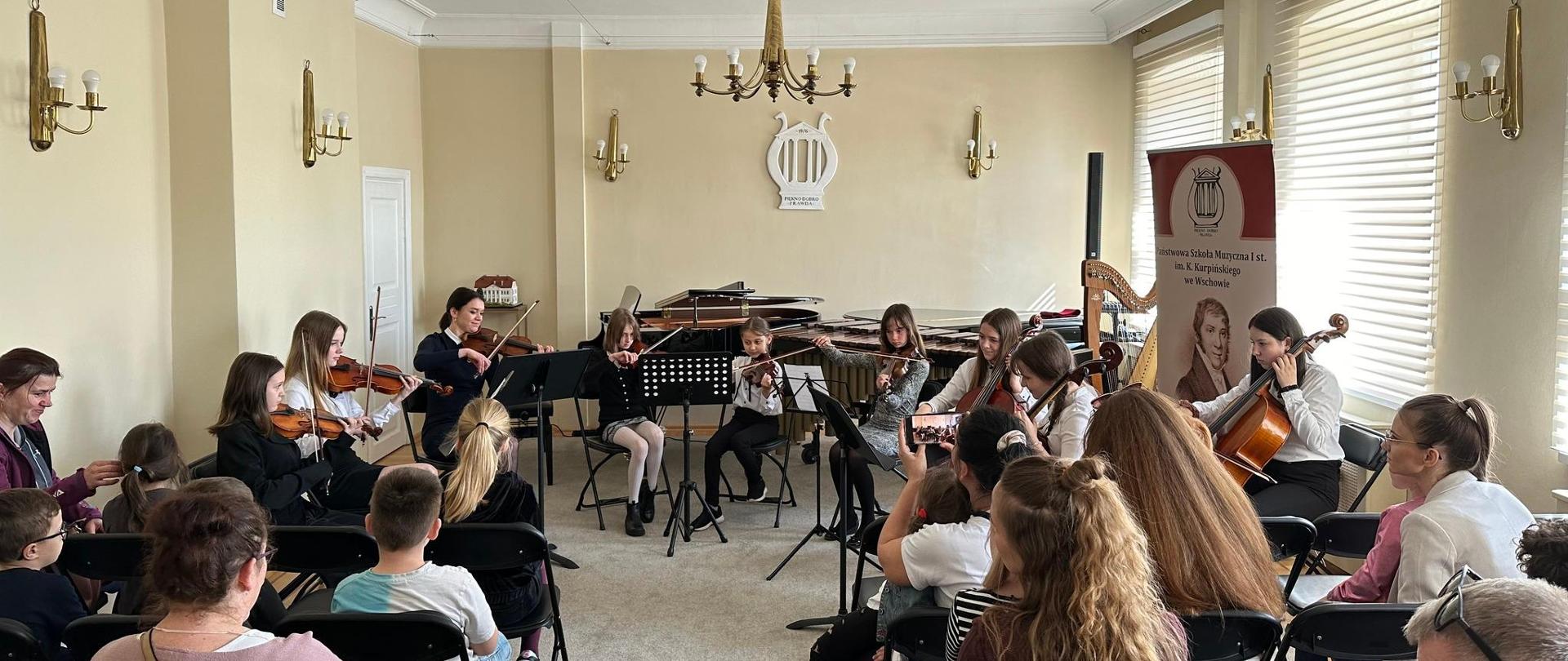 Dziewięcioosobowy zespół smyczkowy występujący w auli szkoły i siedząca tyłem publiczność.