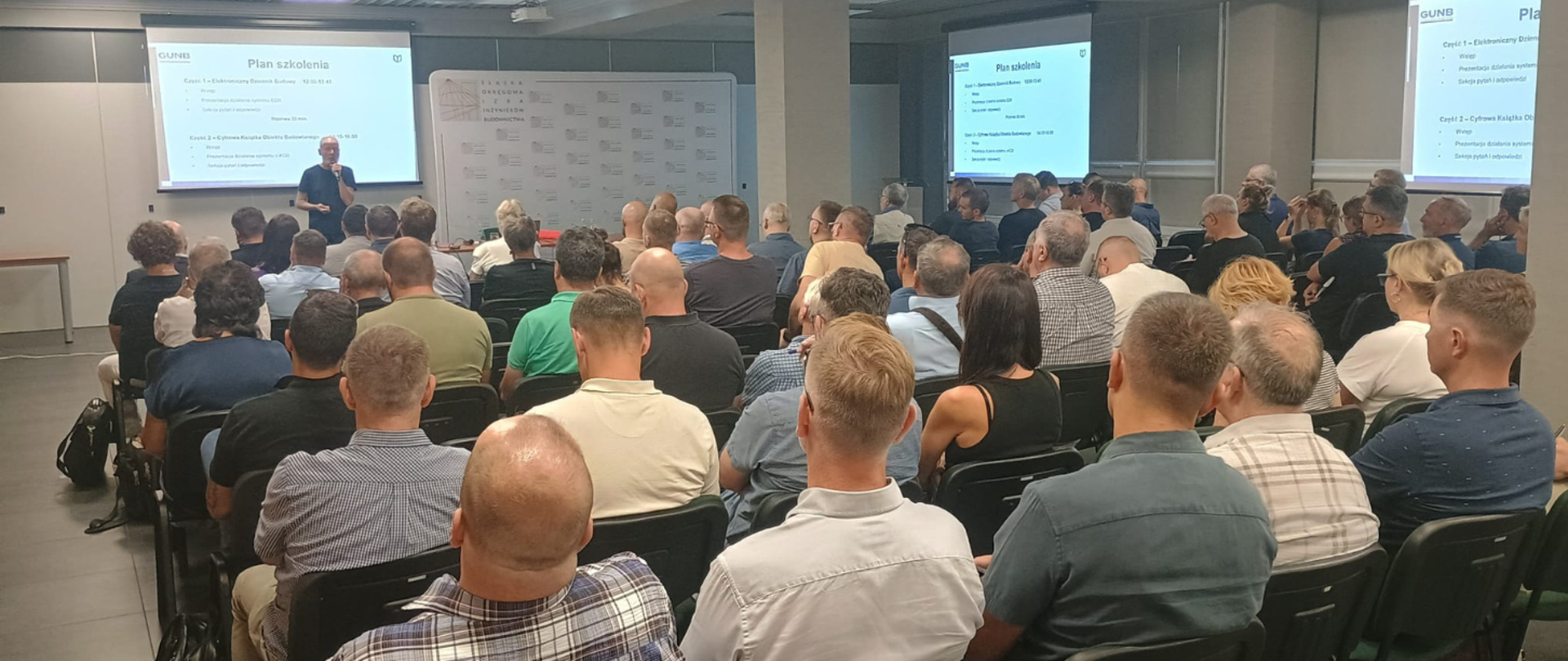 Na zdjęciu sala szkoleniowa – uczestnicy siedzą przy stołach, słuchają prelegenta, na ekranie widoczny jest slajd z planem szkolenia dotyczącym Elektronicznego Dziennika Budowy i Cyfrowej Książki Obiektu Budowlanego.