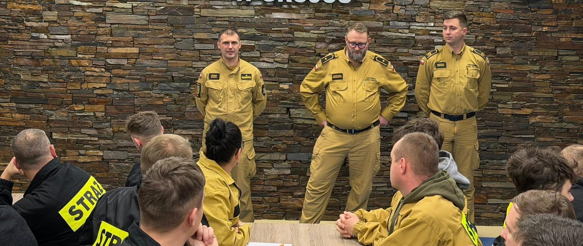 Na zdjęciu widać trzech strażaków stojących przed ścianą z kamienia dekoracyjnego. Mają żółte mundury służbowe.
Jeden z nich przemawia – ma uniesione ręce i wygląda na to, że coś wyjaśnia. Reszta stoi obok, słuchając.
Na ścianie za nimi znajduje się duży biały napis:
„Komenda Powiatowa Państwowej Straży Pożarnej w Grójcu”.
Obok napisu widać dwa herby – jeden związany ze strażą pożarną, drugi z powiatem. Pomieszczenie wygląda na salę konferencyjną w budynku straży pożarnej. Przed nimi, przy stołach, siedzą inni strażacy – widzimy ich od tyłu. Słuchają osób stojących z przodu.
Spotkanie wygląda na oficjalne szkolenie lub odprawę służbową. Wszyscy zachowują powagę i skupienie.