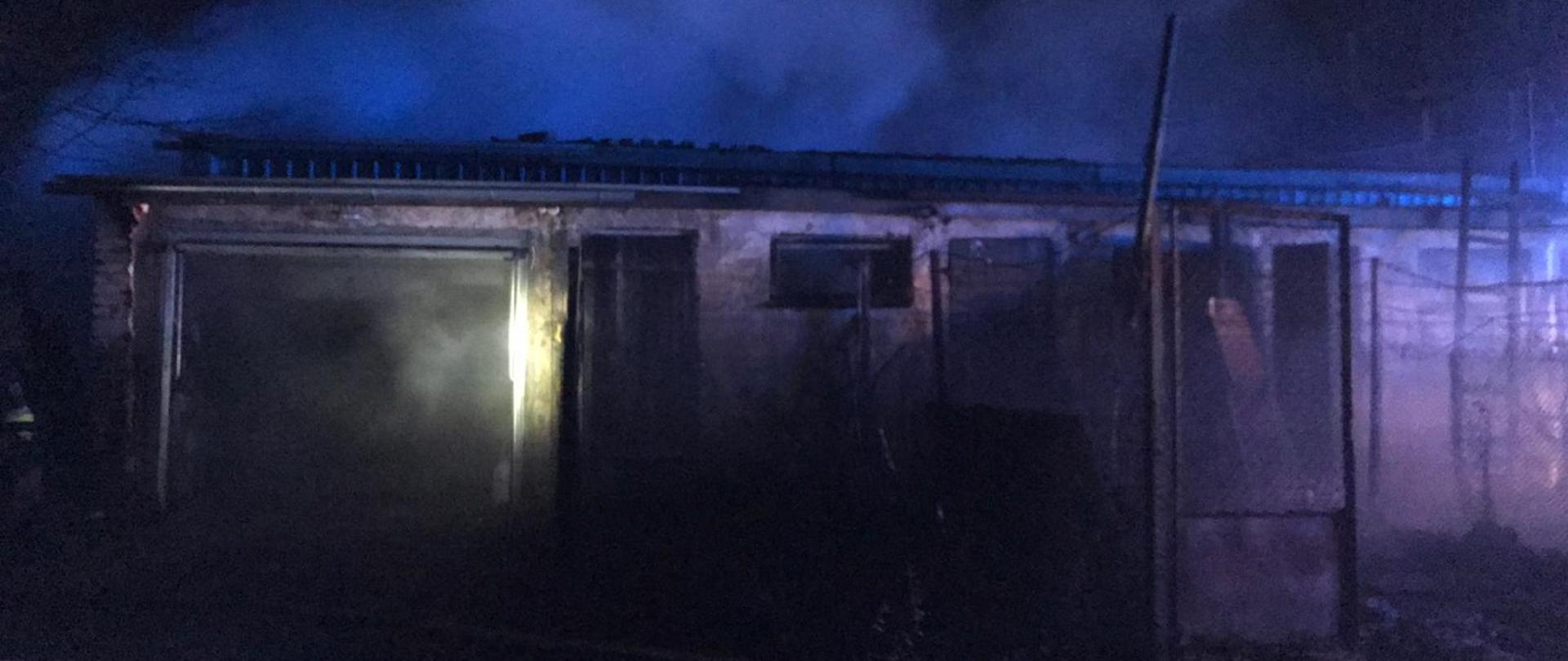 Na zdjęciu widoczny budynek gospodarczy garaż z otartymi drzwiami.Z lewej strony widać strażaka i część samochodu osobowego w biały kolorze.W tle dym. Pora nocna.