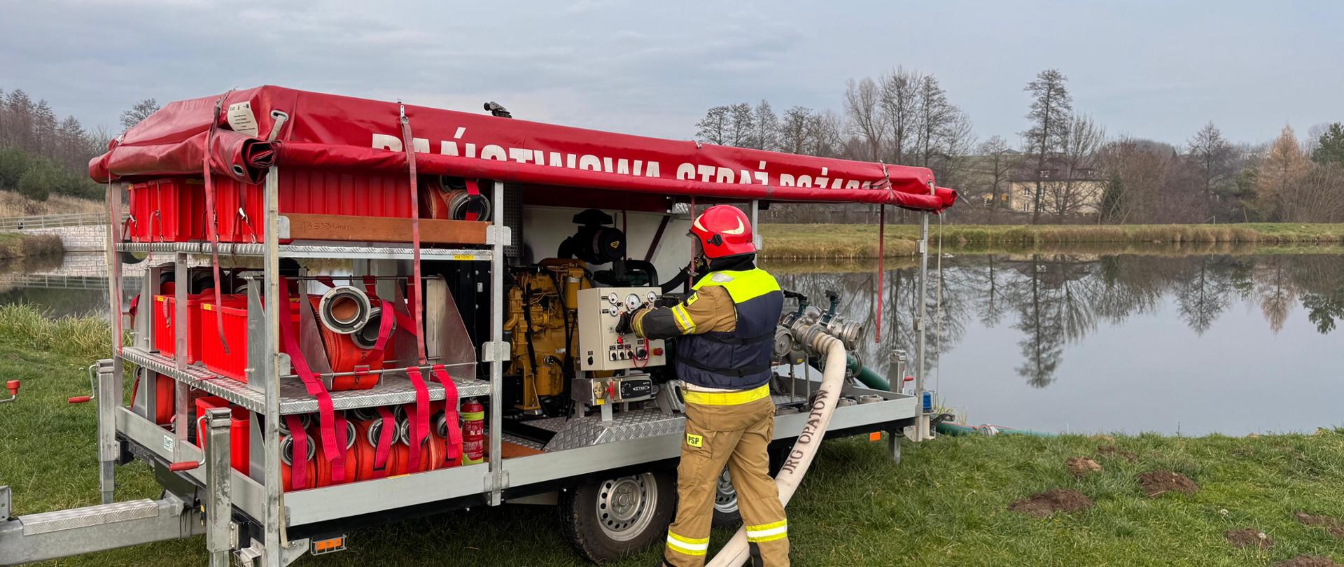Strażak w ubraniu specjalnym przed pompą dużej wydajności. W tle zbiornik wodny