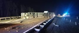 Samochód ciężarowy wraz z naczepą przewrócony na bok blokując autostradę A2 w kierunku Poznania. Uszkodzone barierki. Po drugiej stronie stoi radiowóz. 