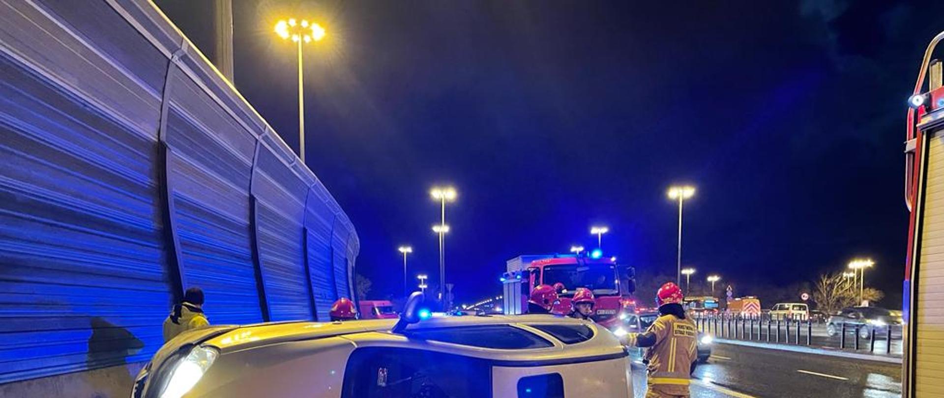 Na pierwszym planie widać leżący na lewym boku samochód osobowy Opel Corsa koloru srebrnego, przy którym stoją strażacy