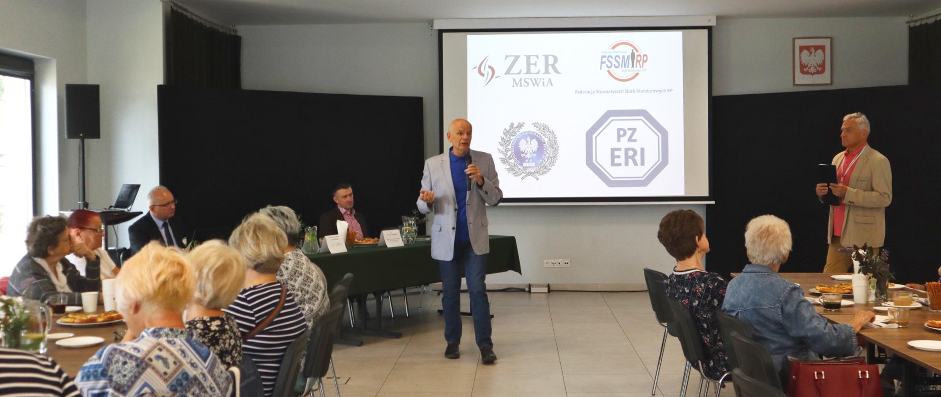 gen. insp. w st. spocz. Leszek Szreder podczas przemówienia, słuchacze siedzą przy stołach, na ścianie wyświetlona prezentacja