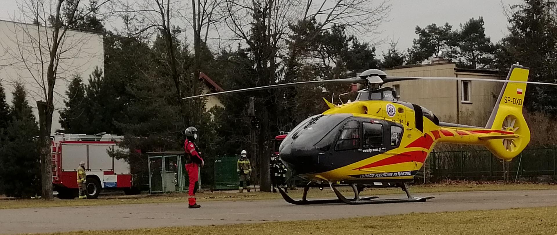 Na zdjęciu widoczny jest czarno-żółty śmigłowiec stojący na betonowym placu przy boisku szkolnym. Maszyna szykuje się do startu. Po lewej stronie widać członka załogi w czerwonym kombinezonie i czarnym hełmie na głowie. Dokonuje on inspekcji śmigłowca przed startem. W tle widać wozy strażackie z włączonymi migającymi światłami alarmowymi. Straż Pożarna zabezpiecza strat maszyny. Za helikopterem widać rząd drzew na których brak jest liści oraz budynki szkoły.
