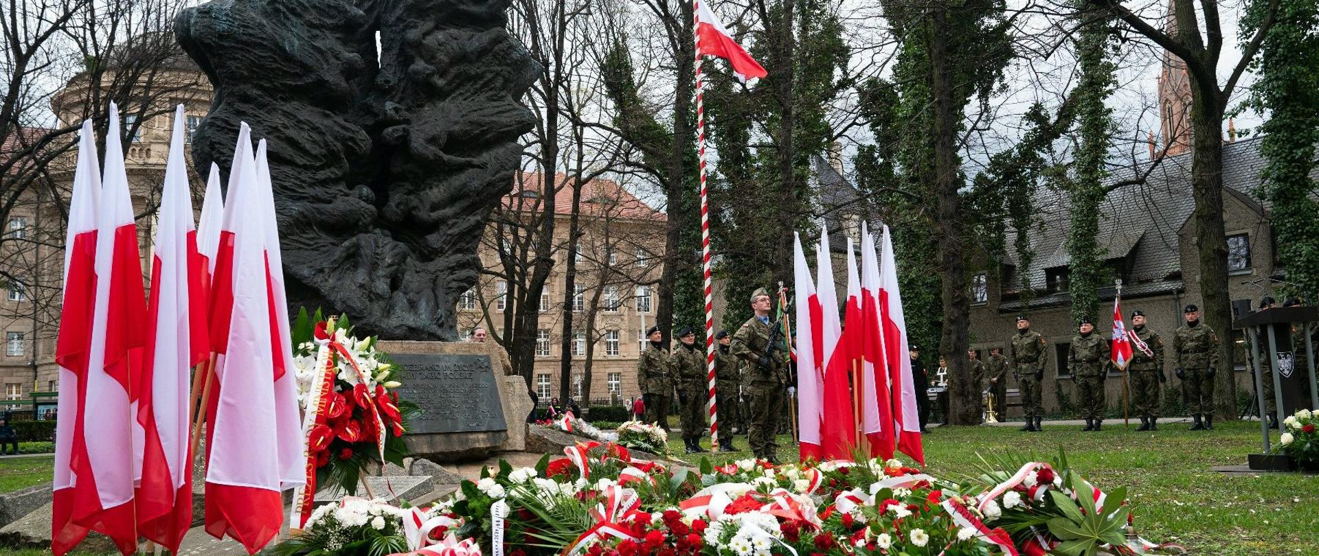 pomnik przed którym leży dużo wieńcy biało czerwonych, z boku stoją flagi biało czerwone w tle żołnierze i budynki