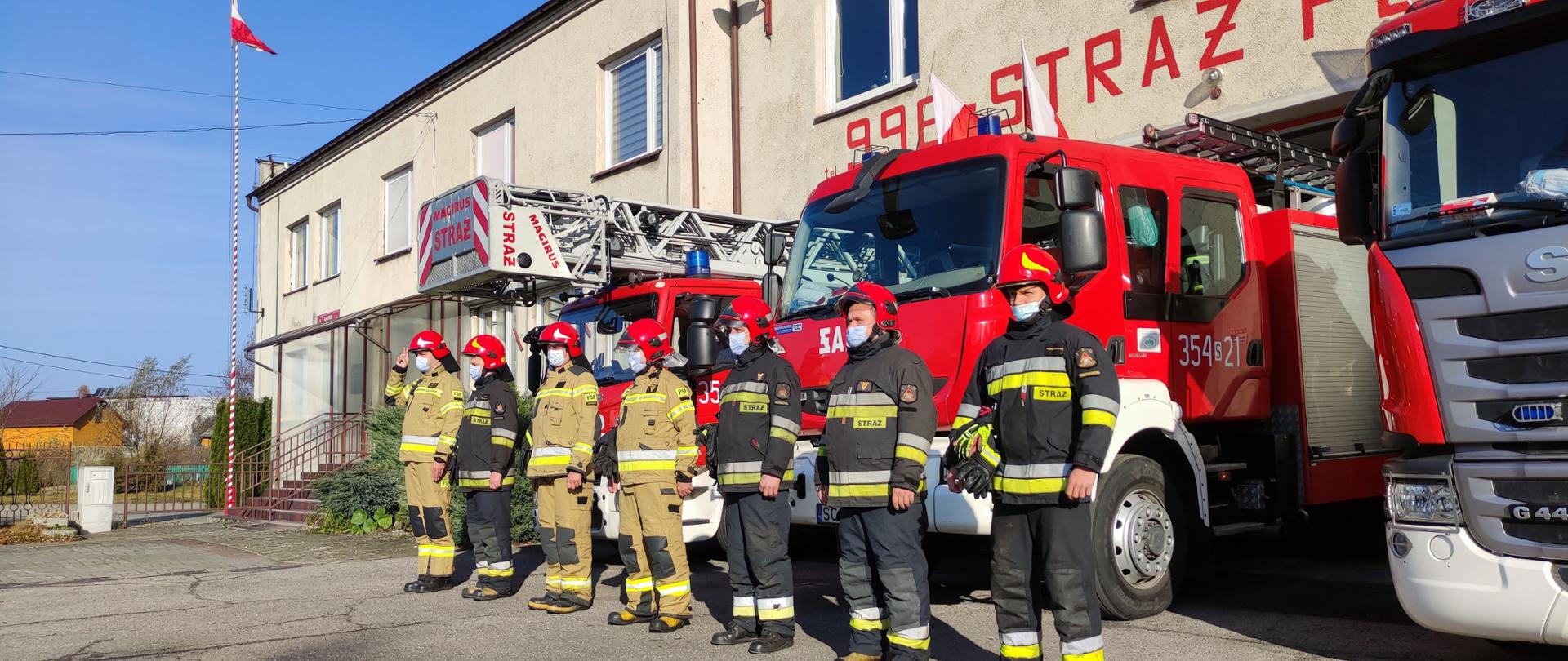 Na zdjęciu widzimy strażaków w umundurowaniu bojowym na tle pojazdów pożarniczych. Na zdjęciu widoczny również maszt z podniesioną Flagą Państwową