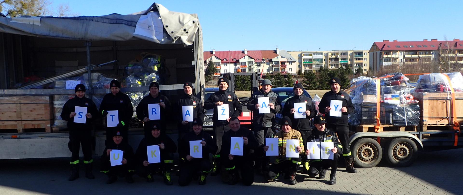 Grupa strażaków stoi z napisem "Strażacy Ukrainie". W tle załadowany na samochody sprzęt pożarniczy.