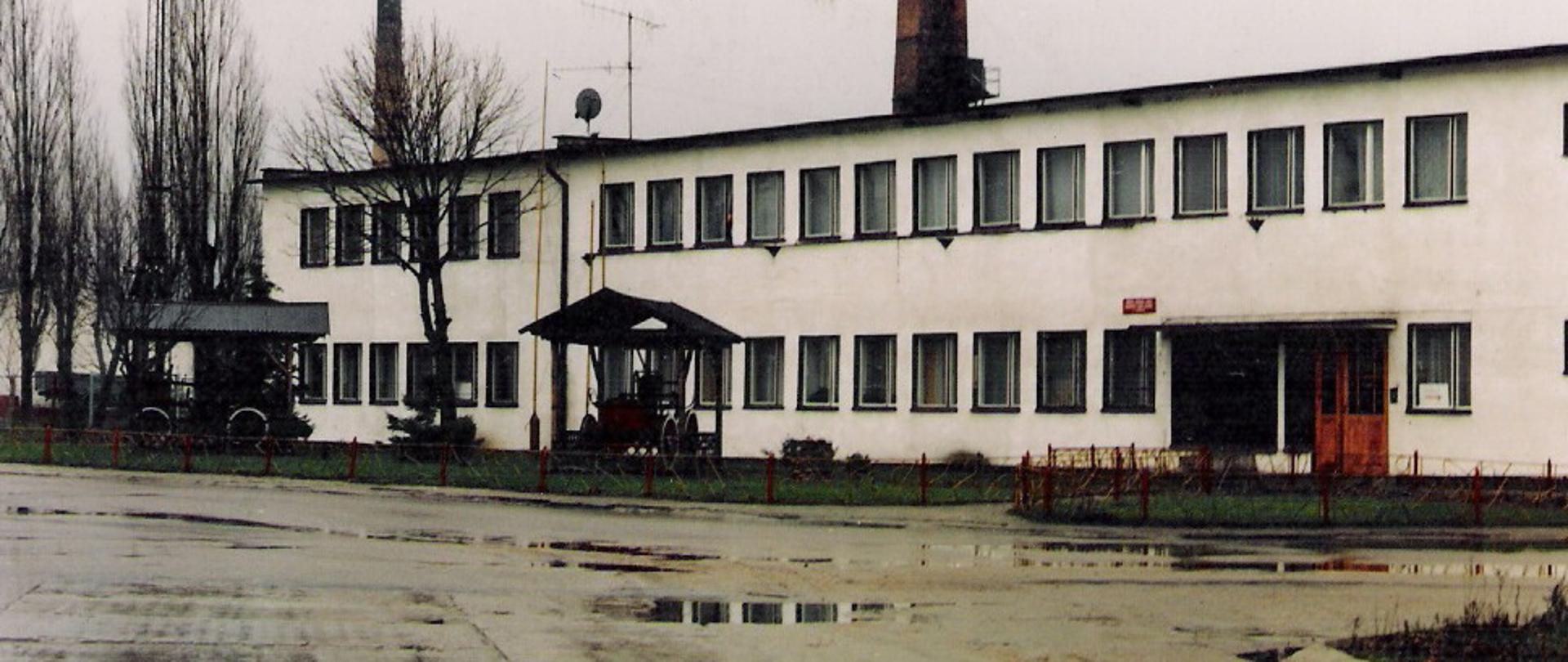 Zdjęcie przedstawia prostokątny szary budynek z licznymi oknami oraz drzwiami wejściowymi. Przed budynkiem stoją dwie sikawki strażackie pod wiatkami, znajdują się również niewielkie drzewa. Za budynkiem widać dwa wysokie kominy wykonane z czerwonej cegły.