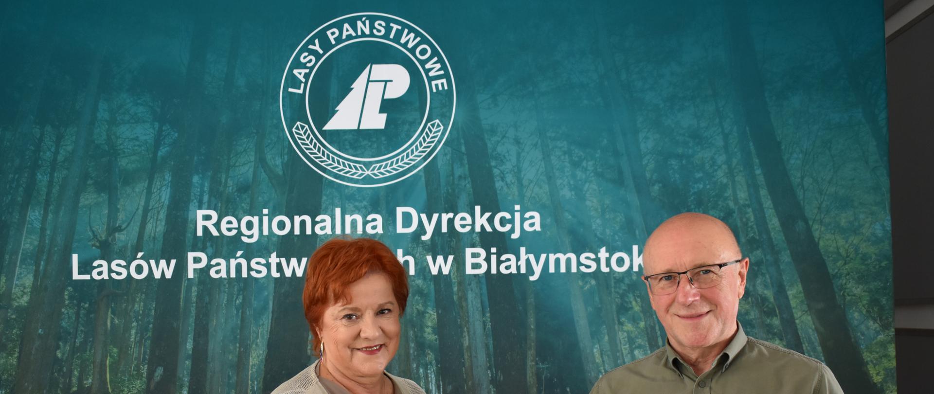 Na zdjęciu znajduje się kobieta i mężczyzna, którzy ściskają sobie dłoń w geście porozumienia. Za nimi znajduje się ścianka promocyjna Regionalnej Dyrekcji Lasów Państwowych w Białymstoku.