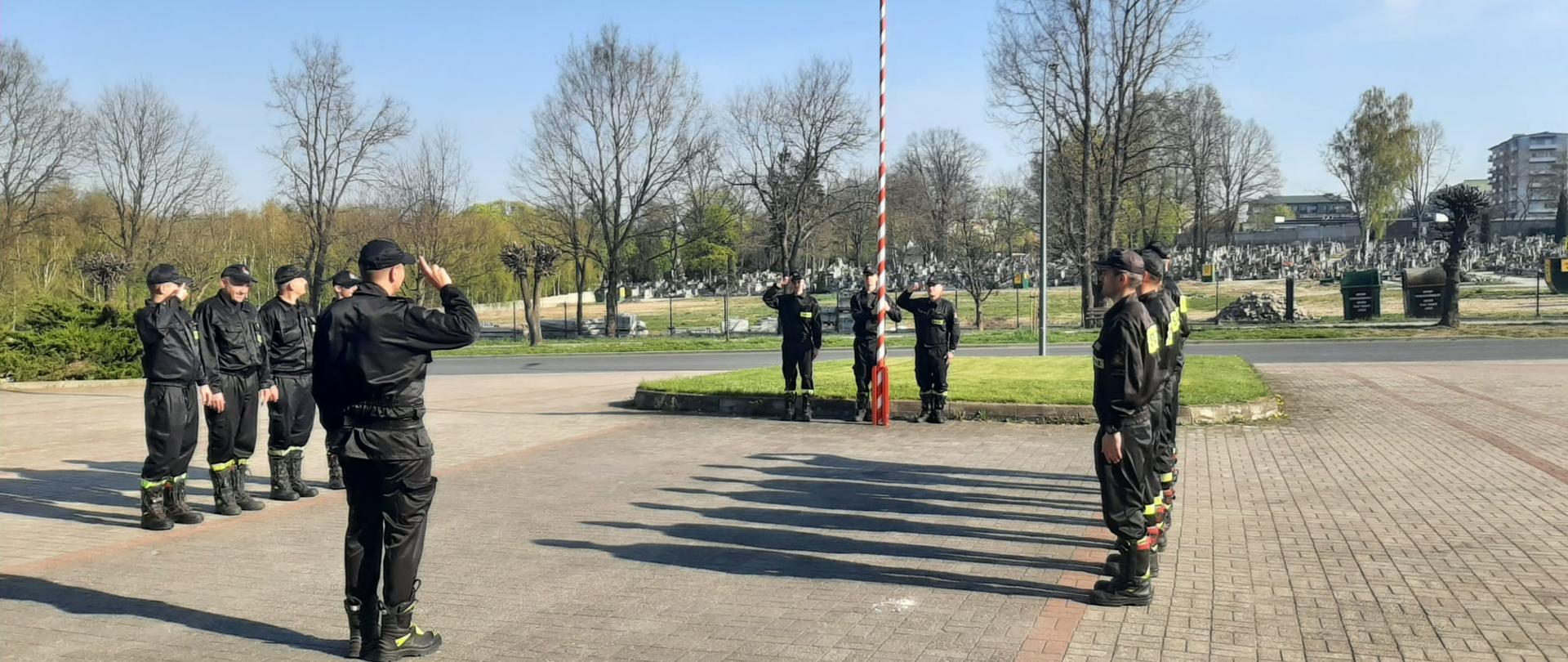 Na zdjęciu widać 18-stu strażaków. 14-stu z nich stoi w dwóch 7-mio osobowych szeregach, zwróconych twarzami do siebie. Między nimi znajduje się strażak (dowódca) zwrócony twarzą do masztu z flagą znajdującym się na przeciw niego. Flagę polski na maszt wciąga 3 strażaków.