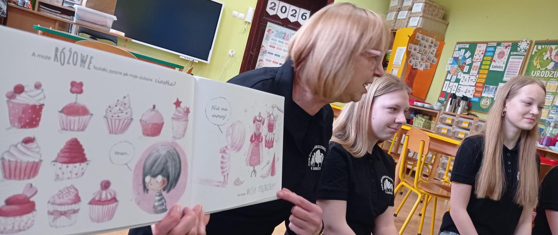 Zbliżenie na otwartą książkę dla dzieci z ilustracjami różowych babeczek i postacią dziewczynki. Książkę trzyma kobieta w czarnej koszuli, wskazując palcem na tekst. Obok siedzą dwie nastolatki w czarnych koszulkach polo, uważnie patrząc w stronę dzieci (poza kadr ). W tle widoczna sala lekcyjna.