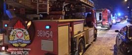 Kolumna wozów straży pożarnej, stojących wieczorową porą na ulicy przed budynkiem wielorodzinnym