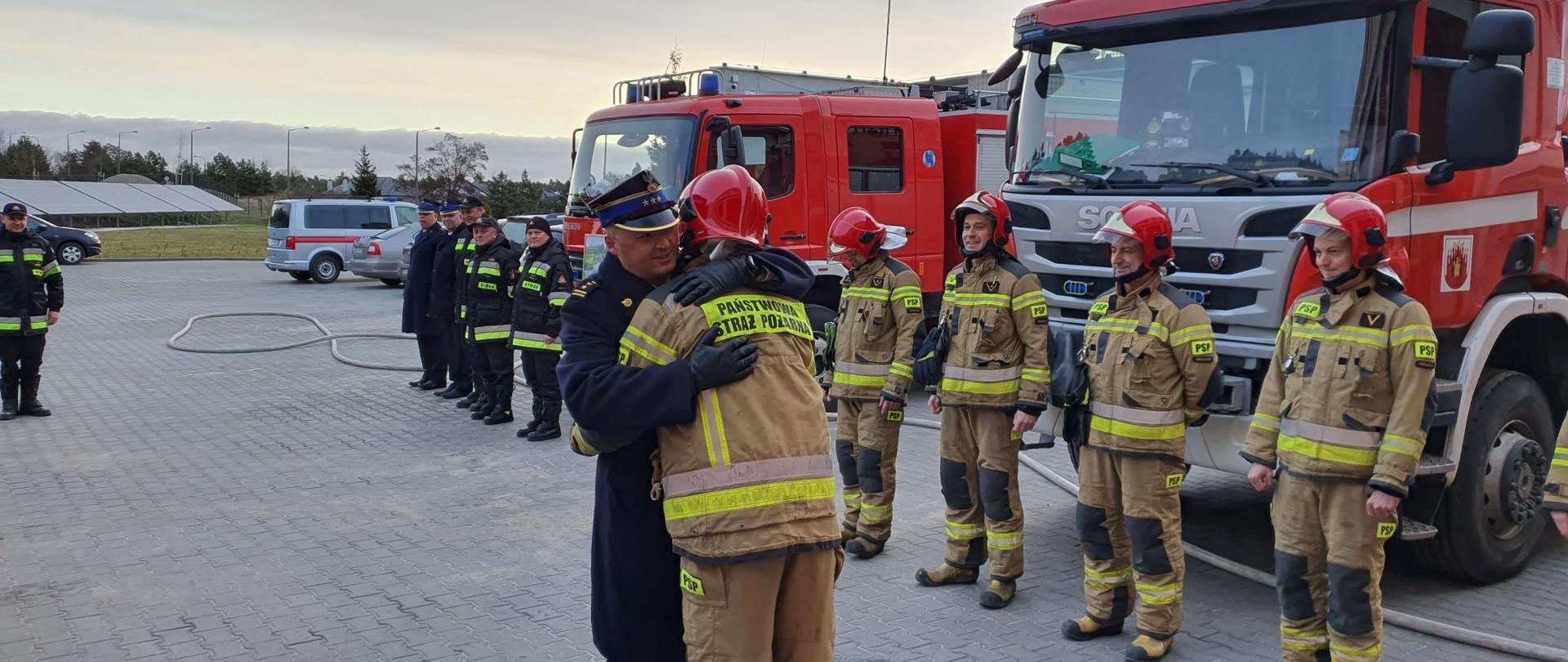 Zdjęcie przedstawia utwardzony plac, na którym odbywa się zbiórka strażaków ubranych w mundury o kolorze piaskowym i czerwone hełmy stojących w szeregu. Pośrodku dwóch strażaków obejmuje się w przyjacielskim uścisku. W tle 2 czerwone wozy bojowe.