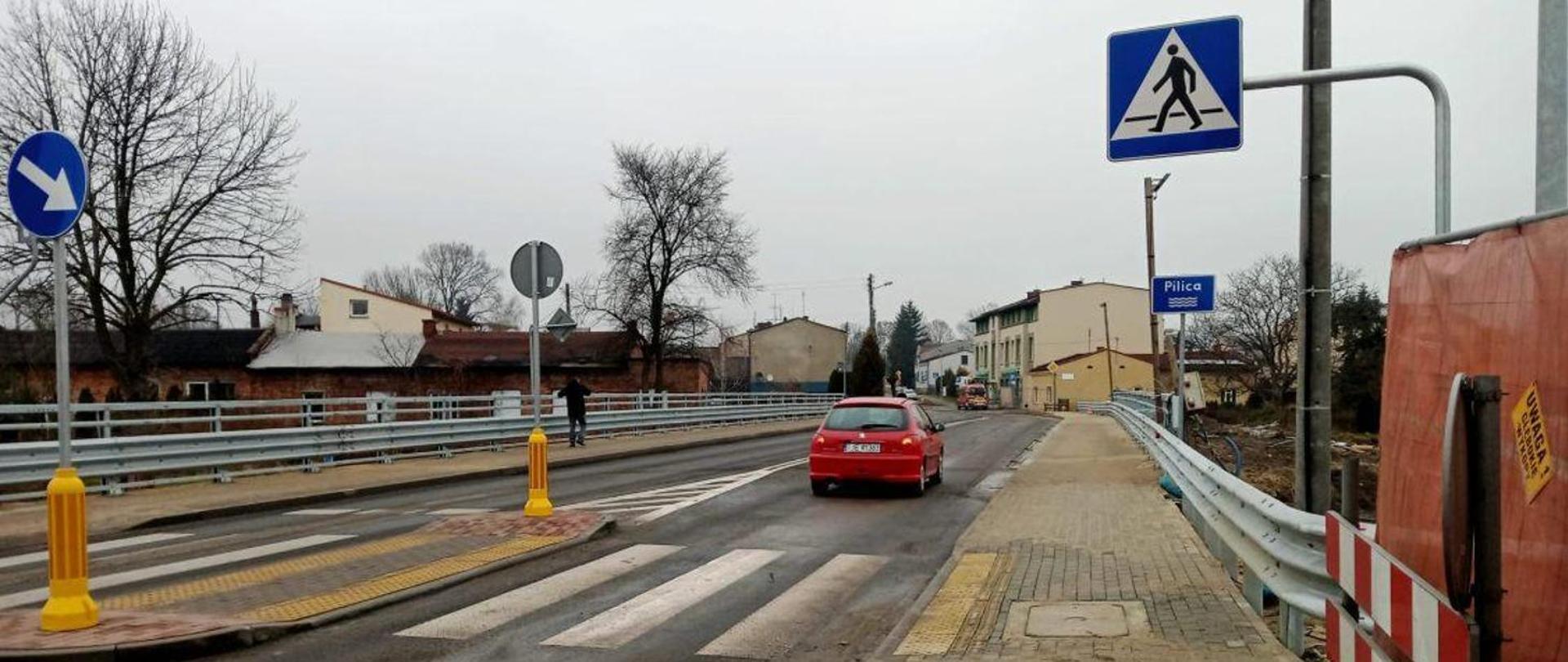 Widok nowo wyremontowanego odcinka drogi z przejściem dla pieszych i wyspą dzielącą pasy ruchu. Na jezdni znajduje się czerwony samochód, a po prawej stronie widoczny jest chodnik z barierkami ochronnymi oraz znak informujący o przejściu dla pieszych. W tle znajdują się zabudowania miejscowości Piłka, a po lewej stronie widoczne są dodatkowe znaki drogowe i barierki zabezpieczające.