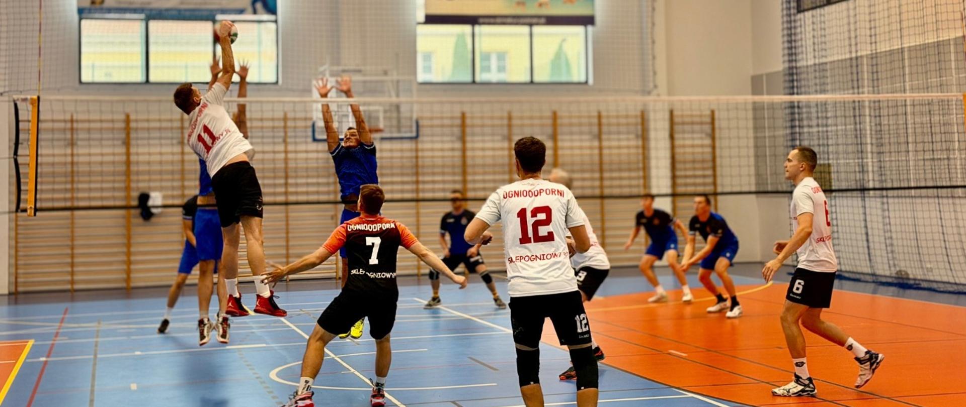 Strażacy grają w piłkę siatkową. Jeden ze strażaków atakuje, dwóch strażaków blokuje 