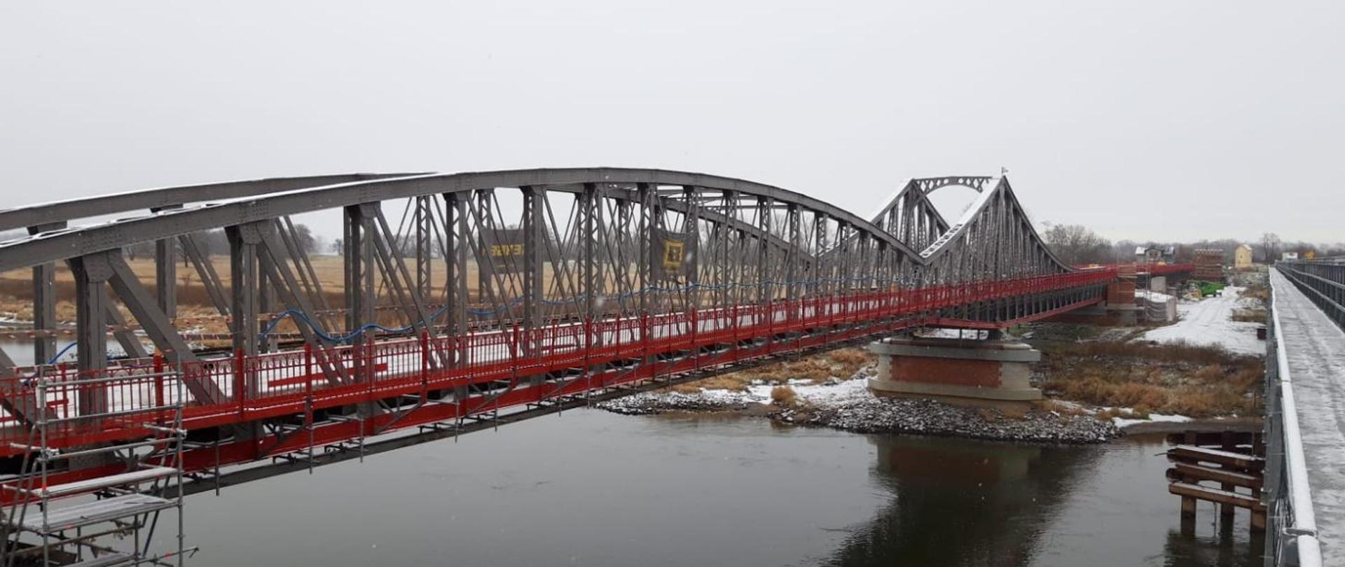 Stalowy most z barierkami w kolorze czerwonym z widocznymi elementami rusztowania związanego z trwającym remontem. Poniżej przepływa Ścinawa. Na moście, brzegu i w tle niewielka warstwa śniegu po niedawnych opadach.