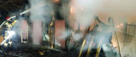 Na zdjęciu strażak walczący z ogniem. Strażak ma czerwony hełm i piaskowe ubranie. W rękach trzyma wąż zakończony prądownica. Gasi palący się budynek gospodarczy.