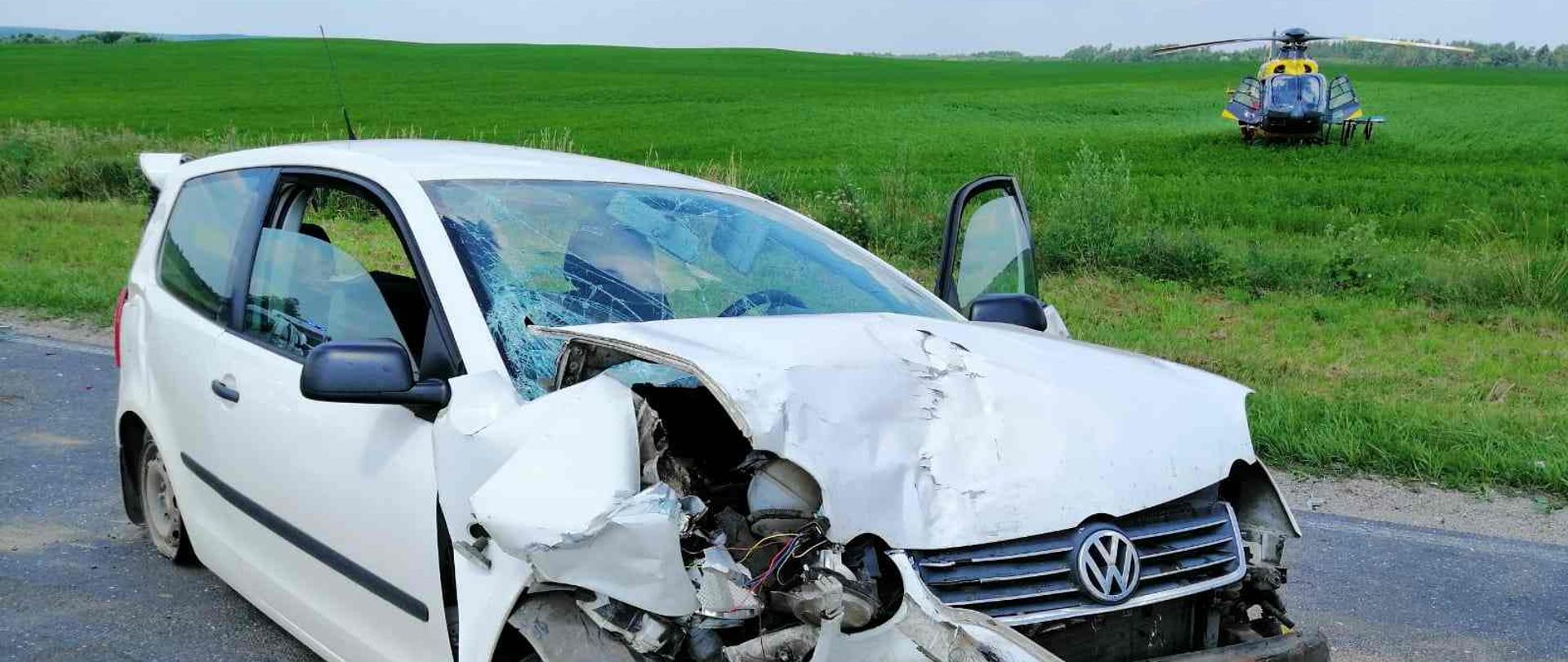 Na pierwszym planie rozbity samochód w tle stojący śmigłowiec pogotowia ratunkowego.