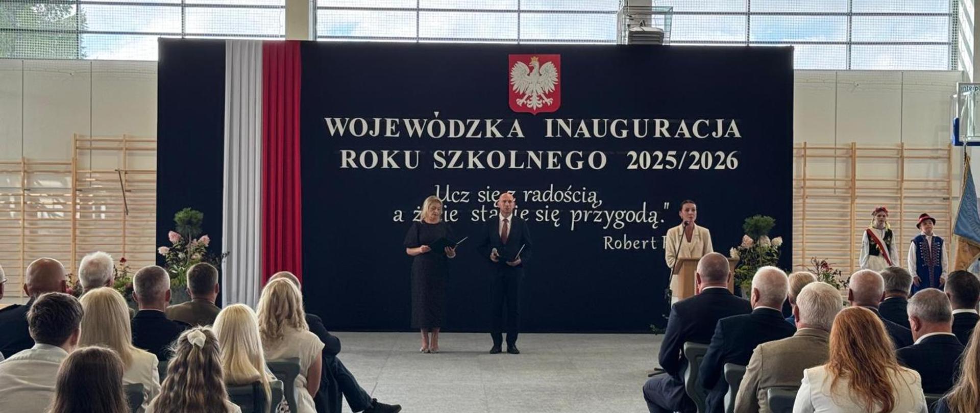 Udekorowana ścianka z okazji wojewódzkiej inauguracji roku szkolnego 2025/2026, przed którą znajdują się osoby prowadzące uroczystość oraz przemawiająca pani wojewoda pomorska.
