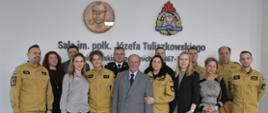 Funkcjonariusze pracownicy cywilni Wydziału Kwatermistrzowskiego mężczyzna w garniturze oraz kierownictwo Komendy Wojewódzkiej Państwowej Straży Pożarnej stoją obok siebie na tle ściany z logiem Państwowej Straży Pożarnej oraz popiersia Tuliszkowskiego na ścianie jest napis sala imieniem podpułkownika Józefa Tuliszkowskiego.