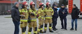 Ćwiczenia w ZSZ. Na parkingu szkolnym stoją organizatorzy i oraz obserwatorzy ćwiczeń. Wśród nich jest 4 strażaków, 2 policjantów oraz dwóch mężczyzn w ubraniach cywilnych. Mężczyźni stoją obok siebie. Z prawej strony widoczny jest fragment wozu strażackiego, a w tle budynki. 