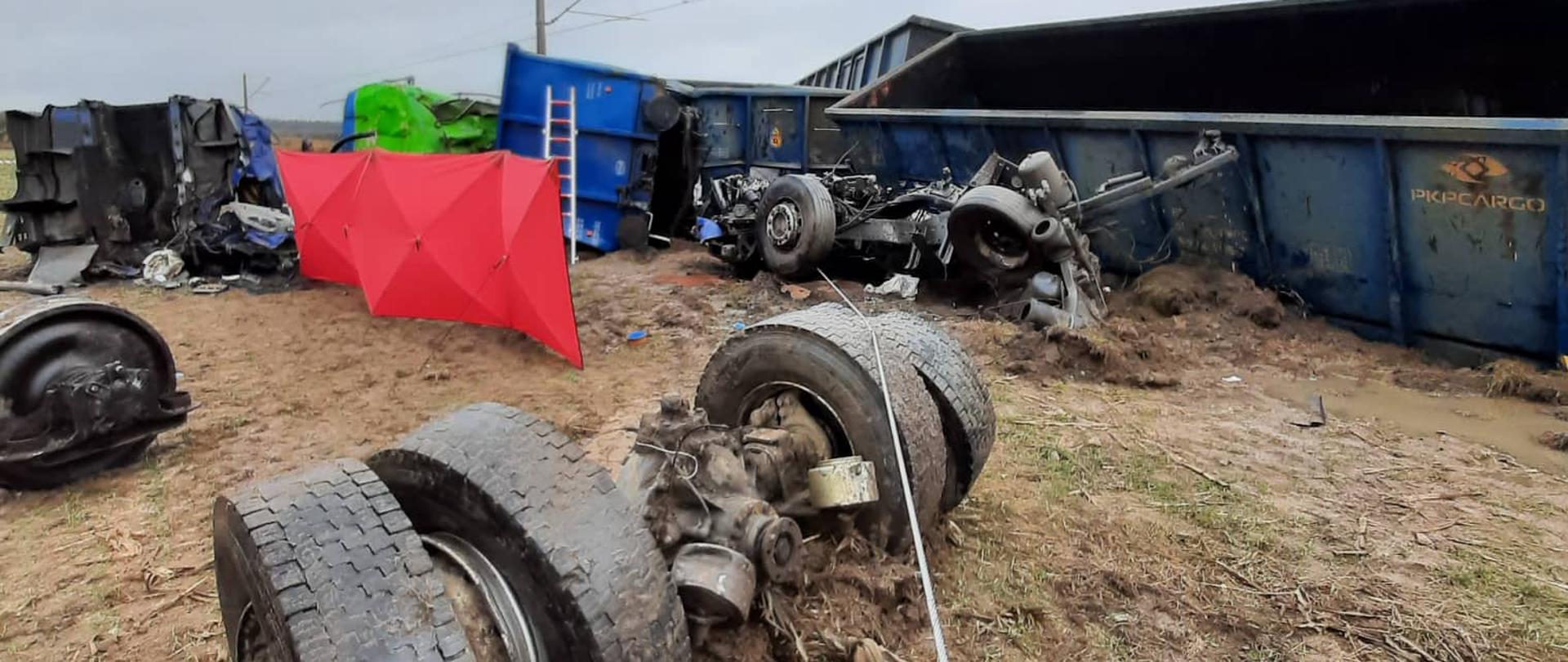 Zdjęcie przedstawia rozczłonowany pojazd typu TIR oraz wykolejone wagony składu kolejowego. Widać także czerwony parawan zasłaniający zwłoki. 