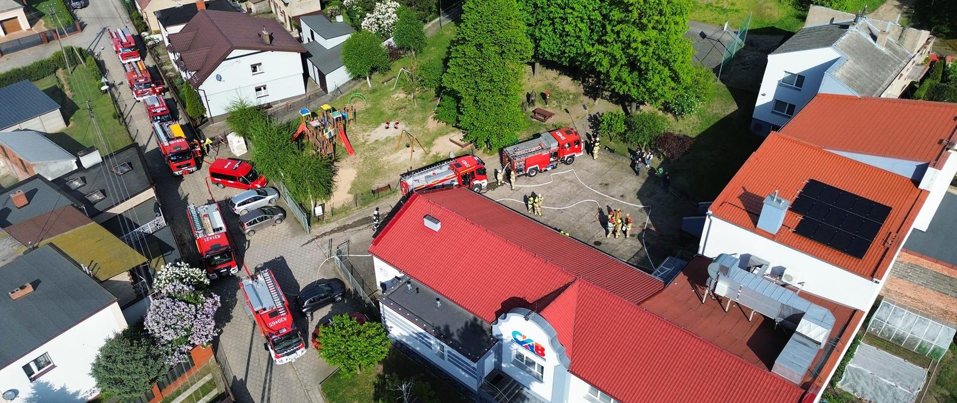 Miejsce ćwiczeń z lotu ptaka. Przy budynku stoją wozy strażackie wzdłuż drogi. Obok wozów chodzą strażacy i druhowie. 