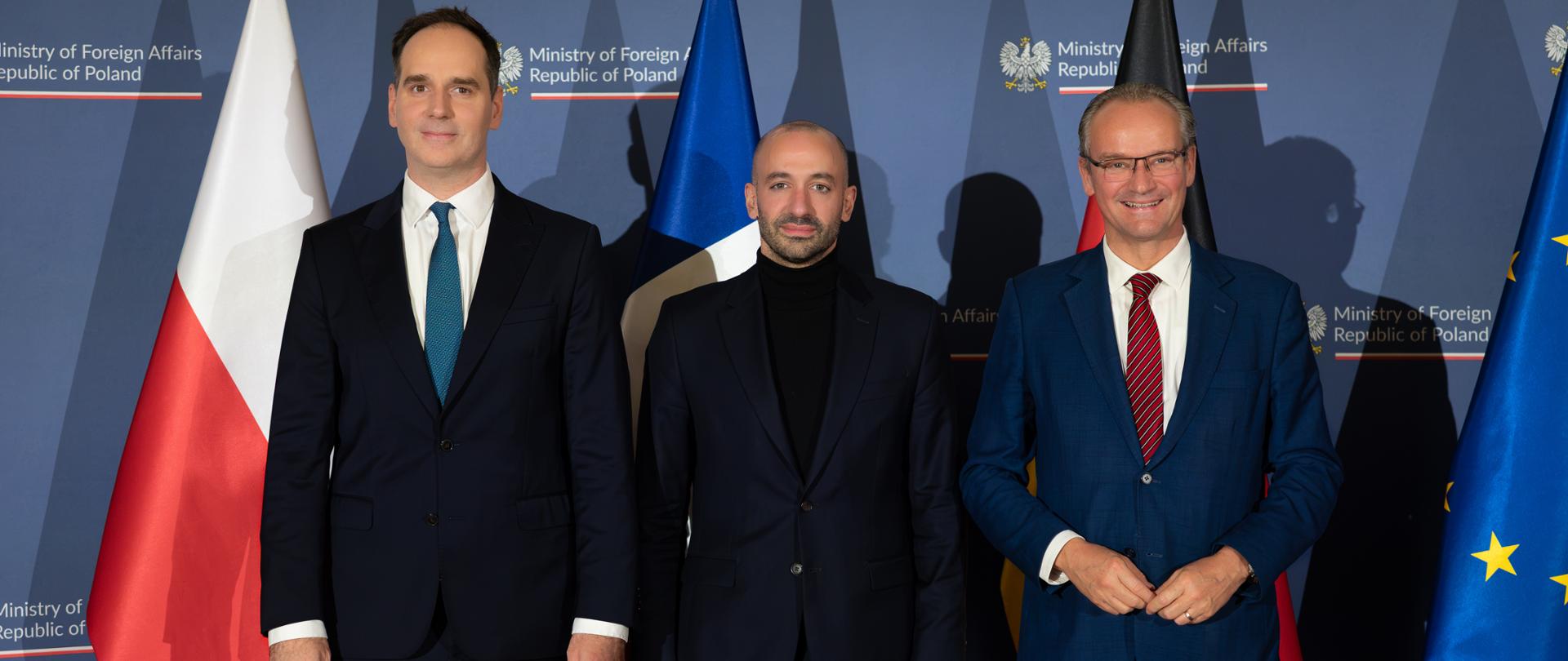 Trzech wiceministrów pozuje do wspólnego zdjęcia na tle flag Polski, Unii Europejskiej, Francji i Niemiec. W tle widoczne jest logo Ministerstwa Spraw Zagranicznych RP.