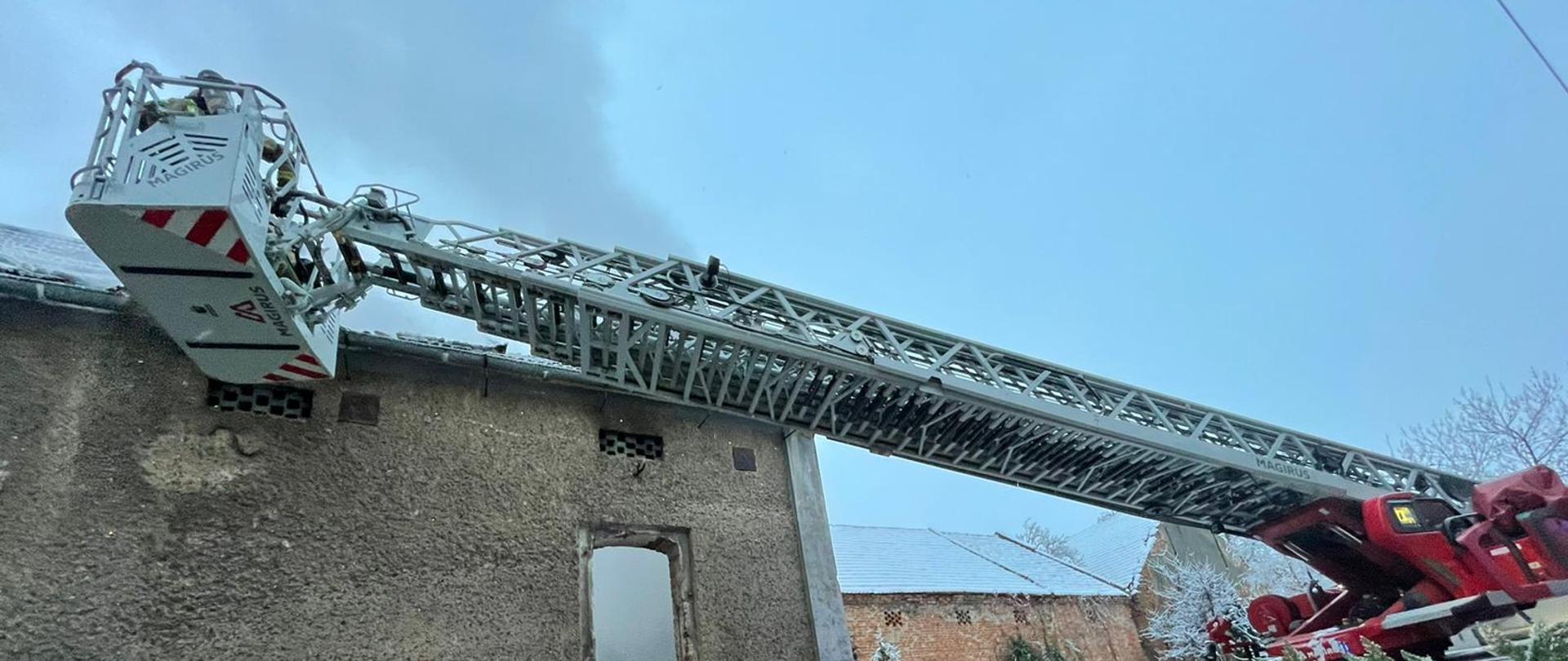 Zdjęcie przedstawia budynek gospodarczy podczas pożaru. Przy elewacji budynku widoczny kosz ratowniczy drabiny mechanicznej podczas działań gaśniczych a w prawej części kadru widoczna reszta drabiny mechanicznej. Powyżej dachu budynku widoczny szary dym z pożaru.