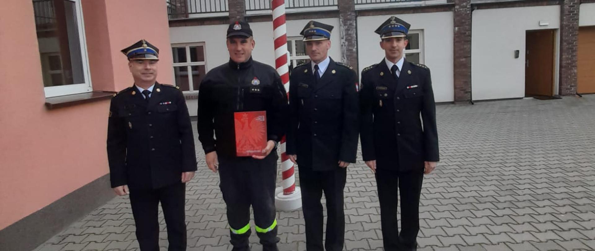 Na zdjęciu można zauważyć czterech strażaków, pośrodku stoi wyróżniony strażak trzymający czerwoną teczkę z nagrodą.