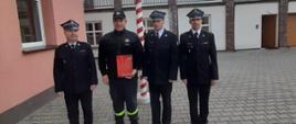 Na zdjęciu można zauważyć czterech strażaków, pośrodku stoi wyróżniony strażak trzymający czerwoną teczkę z nagrodą.