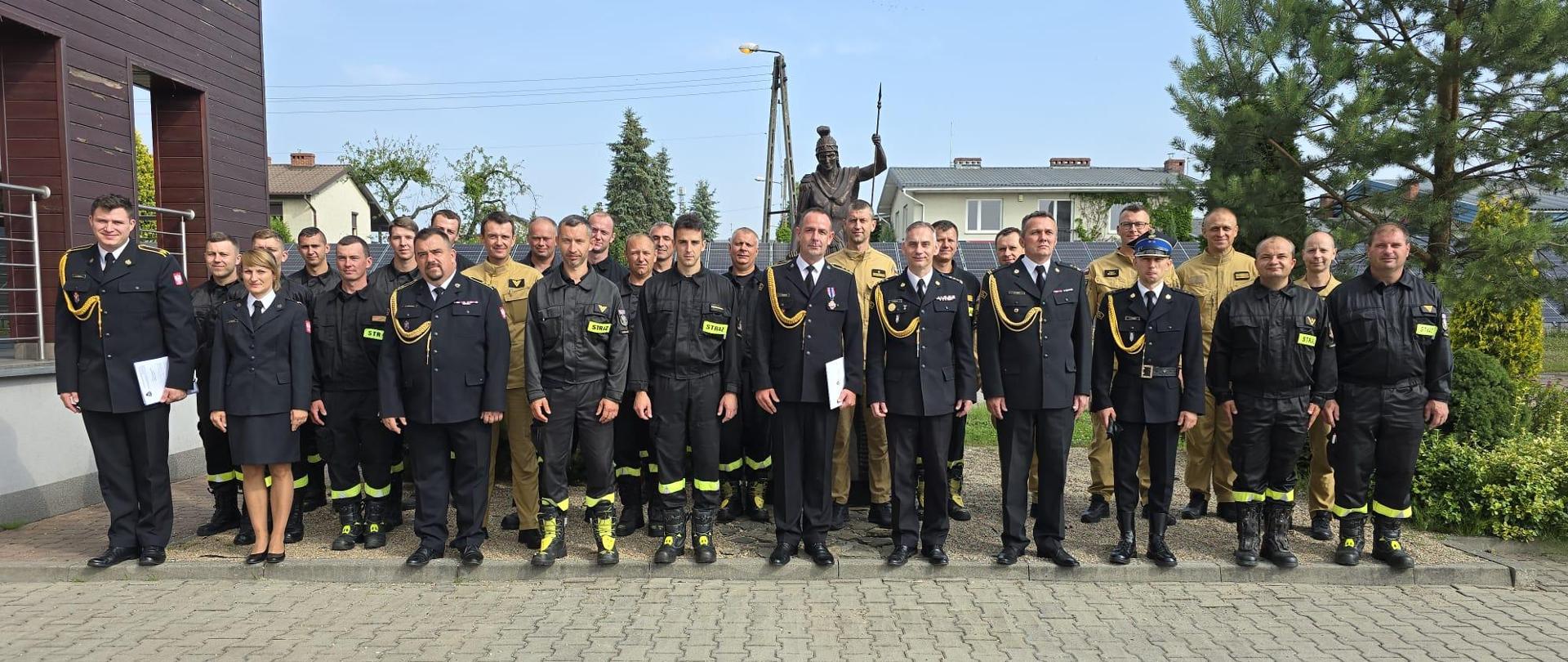 Zdjęcie zrobione na tle figurki św. Floriana. Na pierwszym planie pozuje grupa strażaków w umundurowaniu galowym i koszarowym.