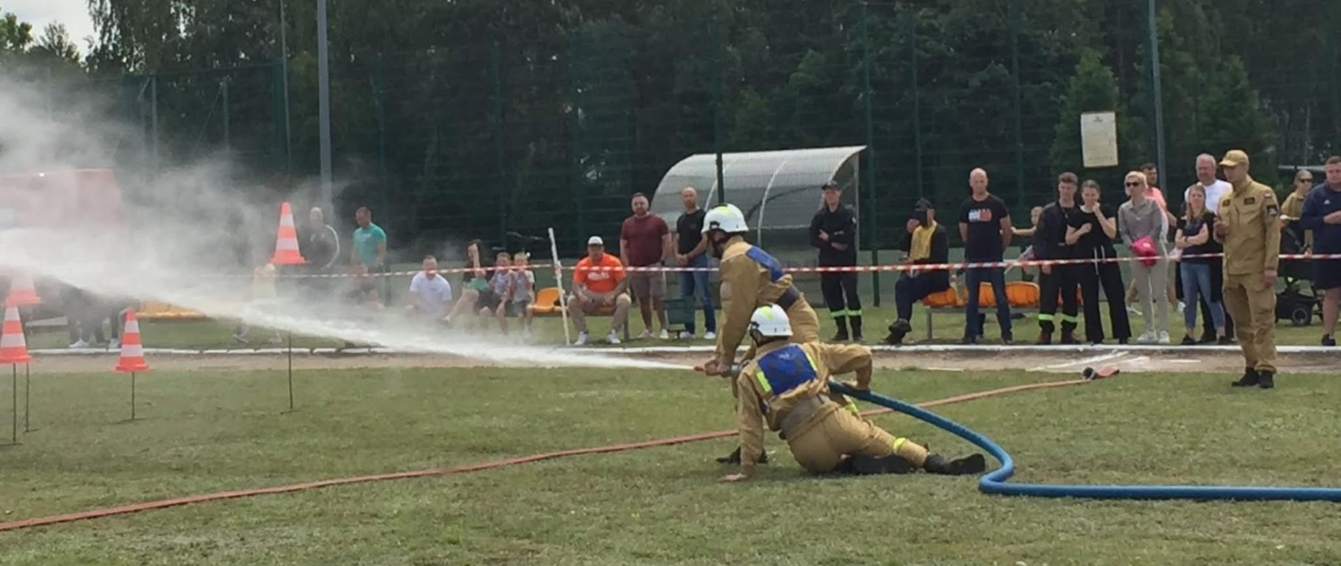 Zdjęcie przedstawia dwóch strażaków OSP ubranych w piaskowe mundury i białe hełmy. Strażacy podają prąd wody w celu strącenia pachołków. W tle widać widzów ora sędziego mierzącego czas ćwiczenia bojowego.