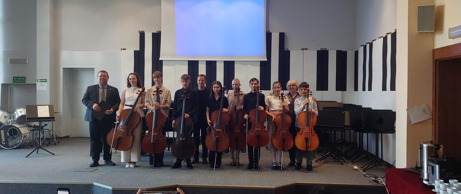 Kobieta i dwóch mężczyzn stoją na scenie ze wszystkimi wykonawcami trzymającymi swoje wiolonczele.