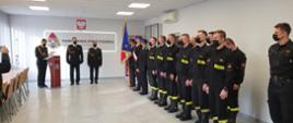 Grupa strażaków w umundurowaniu koszarowym stojąca na zbiórce w sali. Na wprost trzech strażaków w ubraniu galowym. Po prawej stronie jeden ze strażaków trzyma sztandar komendy. W tle baner z logo PSP. Nad nim godło Polski. 