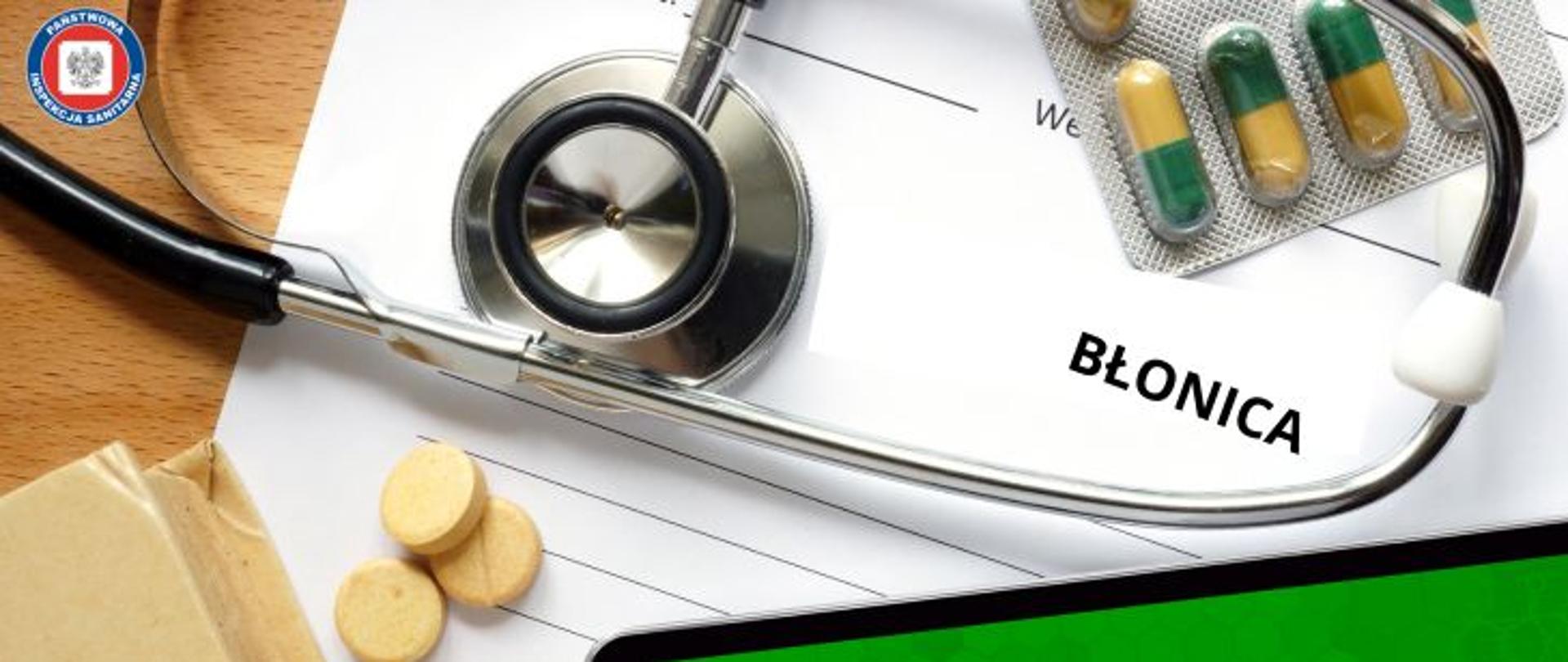 Na stole biała karta pacjenta z czarnym napisem Błonica. Wokół stetoskop, okrągłe żółte tabletki oraz blister z kapsułkami w kolorze pomarańczowo - zielonym. w lewym górnym rogu okrągłe logo Państwowej Inspekcji Sanitarnej przedstawiające czarnego orła na białym kwadratowym tle, które otoczone jest czerwonym wypełnieniem. Zewnętrzną część logo otacza granatowy okrąg z napisem Państwowa Inspekcja Sanitarna w kolorze białym.