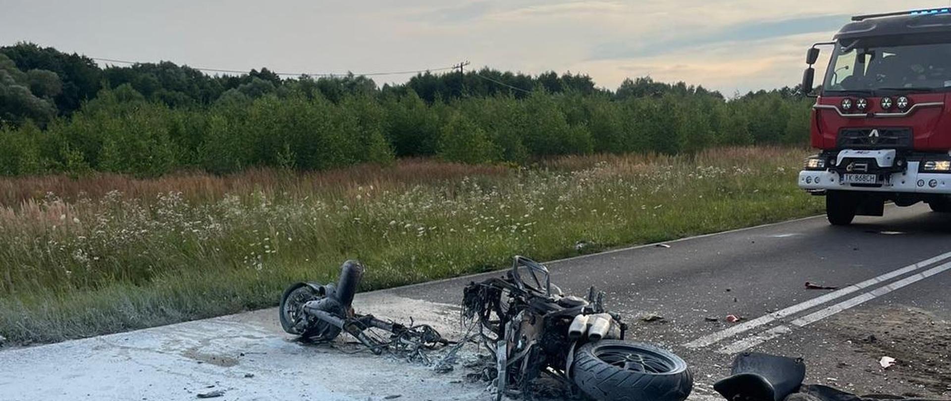 Na jezdni leżący rozwalony motocykl, w tle zieleń na prawo od motocykla widać przód samochodu pożarniczego 