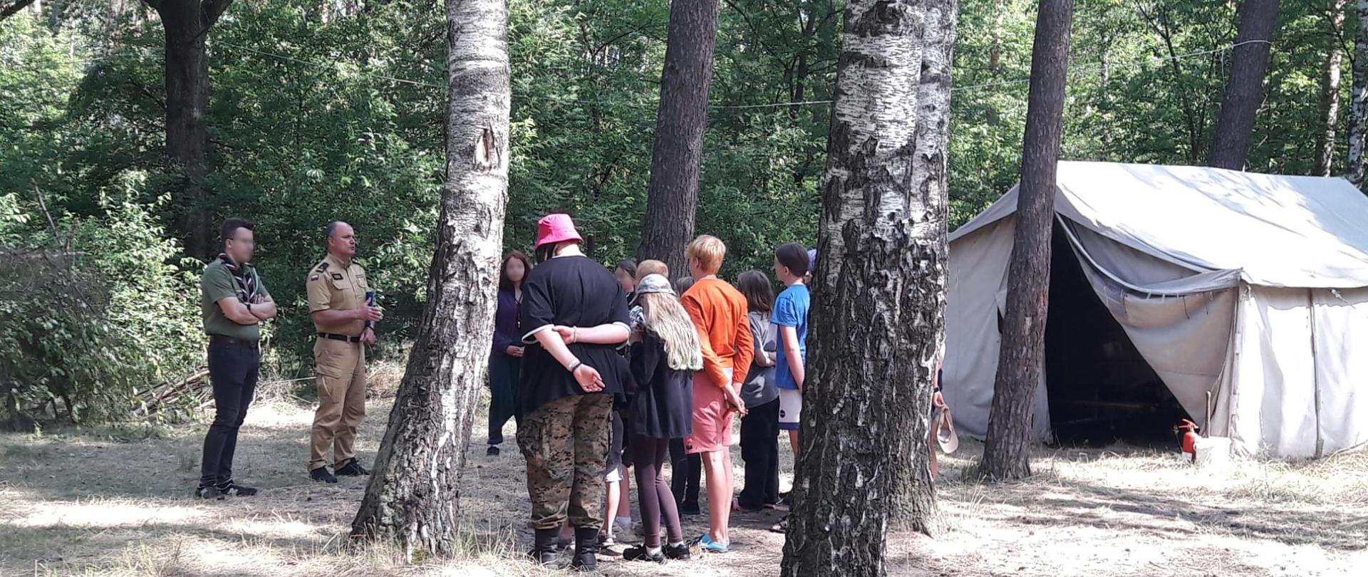 Na zdjęciu funkcjonariusz w ubraniu służbowym w trakcie pogadanki na terenie obozu. Stoi naprzeciw grupy harcerzy. W tle las i namiot.
