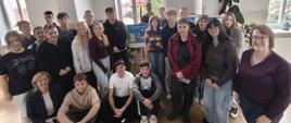 "Zdjęcie przedstawia grupową fotografię uczniów i nauczycielek wykonaną w jasnym, szkolnym korytarzu. Wszyscy są zwróceni w stronę obiektywu i uśmiechają się.
Oto szczegółowy opis elementów widocznych na zdjęciu:
Kompozycja i osoby
• Główna grupa: W centrum i po lewej stronie kadru stoi oraz siedzi grupa około dwudziestu nastolatków (dziewcząt i chłopców) w wieku licealnym. Są ubrani w codzienne, młodzieżowe stroje – bluzy, koszulki, jeansy i swetry w stonowanych kolorach.
• Osoby siedzące na przodzie: W pierwszym rzędzie, bezpośrednio na podłodze, siedzą cztery osoby. Od lewej: dojrzała kobieta o blond włosach w eleganckiej, ciemnej marynarce (prawdopodobnie nauczycielka) oraz trzech chłopaków.
• Osoby po prawej stronie: Po prawej stronie zdjęcia, nieco bliżej przodu, stoi dorosła kobieta w okularach, o krótkich, ciemnych włosach, ubrana w bordowy sweter i jeansy. Obok niej stoją trzy dziewczyny w luźnych ubraniach i okularach.
Tło i otoczenie
• Wnętrze: Grupa znajduje się w szkolnym korytarzu o białych ścianach i jasnej, drewnianej podłodze.
• Okna: W tle widoczne jest duże, wysokie okno z białymi ramami, przez które wpada naturalne światło. Po prawej stronie kadru fragment kolejnego okna, na którego parapecie stoi mała roślina doniczkowa.
• Elementy dekoracyjne:
• W głębi, za plecami uczniów, stoi niewielka tablica lub plakat z niebieskim tłem i napisami (prawdopodobnie gazetka szkolna).
• Po prawej stronie, za grupą dziewcząt, stoi duża roślina doniczkowa (monstera lub fikus) o rozłożystych, zielonych liściach.
Atmosfera na zdjęciu jest radosna, swobodna i pamiątkowa."