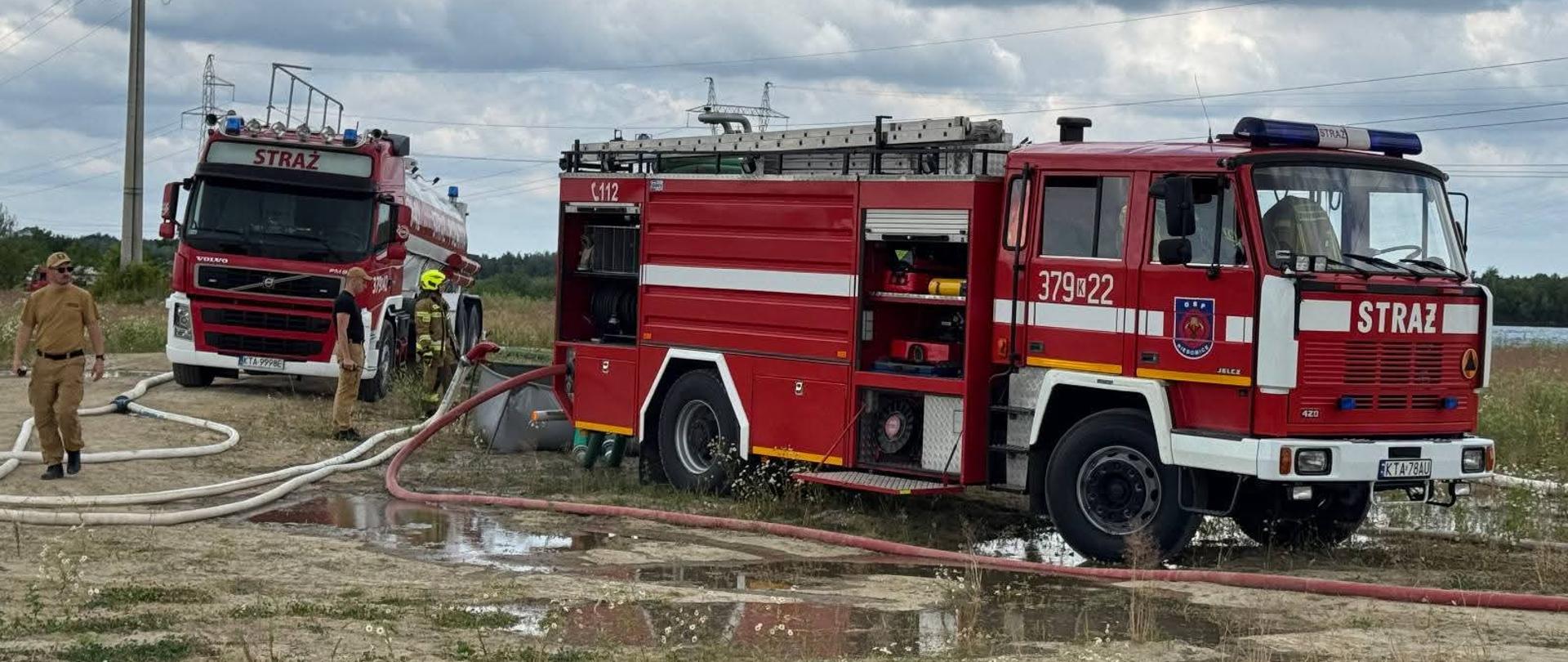 Trzech strażaków w hełmach i ubraniach ochronnych pracuje przy brzegu zbiornika wodnego. Jeden z nich stoi na platformie pojazdu ze sprzętem pożarniczym, drugi trzyma linę, a trzeci wchodzi do wody z linią ssawną. W tle szuwary i niebo z chmurami.